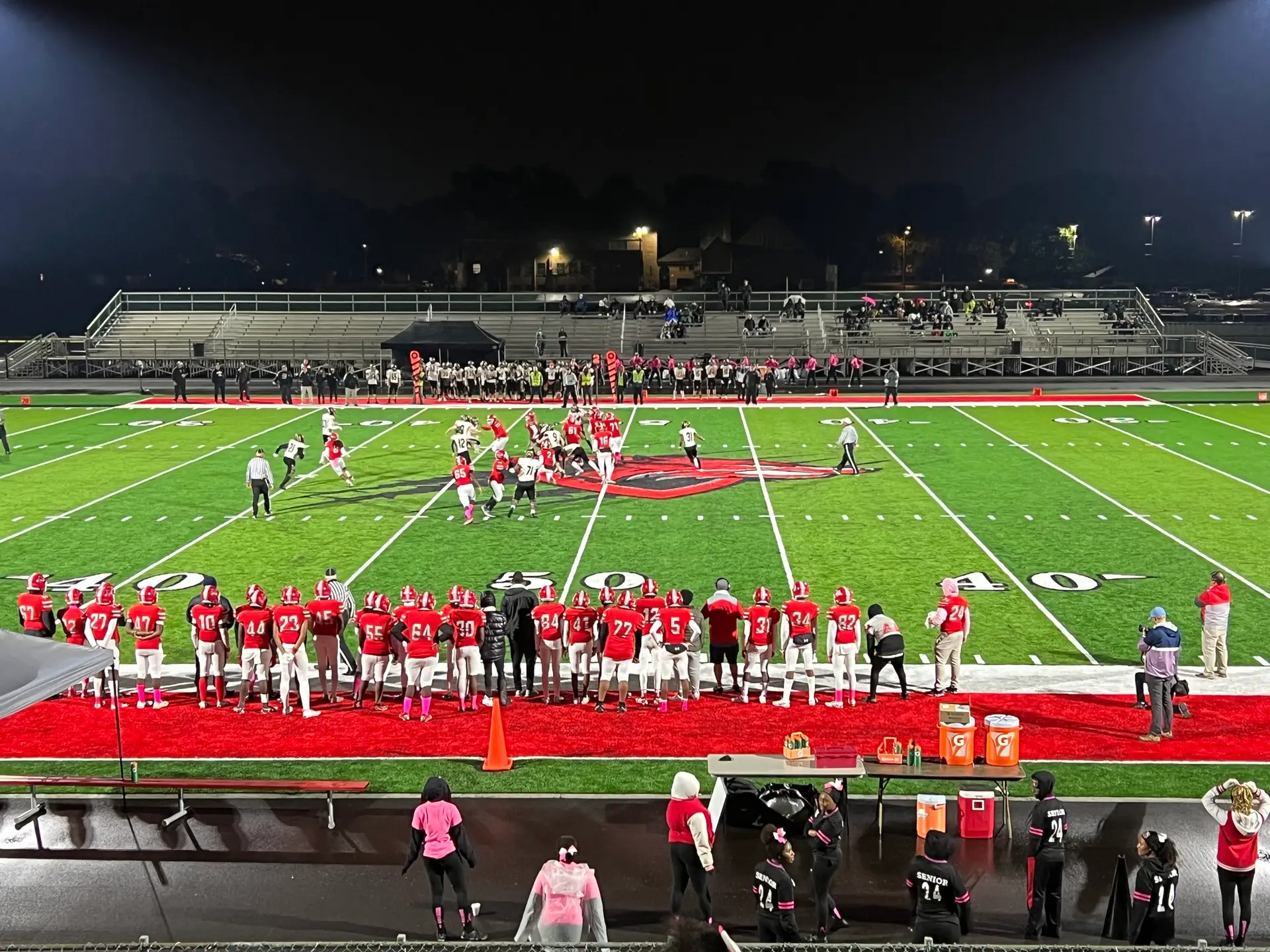 High school football game with players in red uniforms on a green field at night